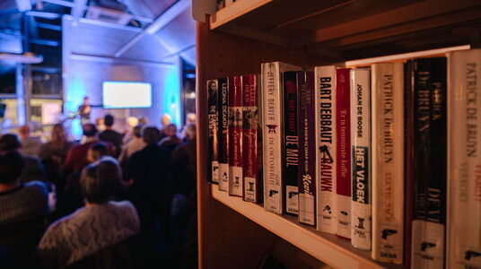 Blik op boekenrek en erachter een gevulde zaal voor lezing